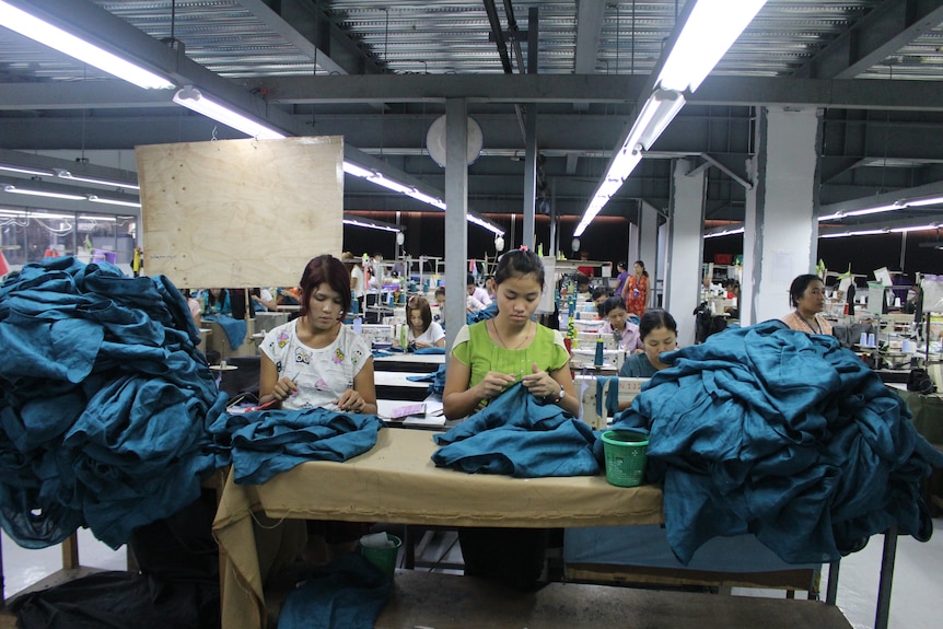 Two women sew by hand garments next to a huge pile of blue fabric.