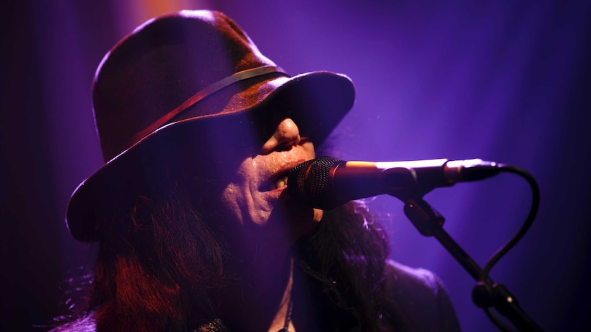 Rodriguez sings into a microphone with a hat covering his eyes
