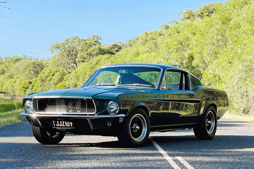 A green Ford Mustang GT with black and gold number plates parked on a road with blue sky and trees.