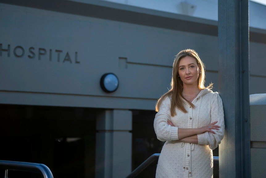 A woman wearing a white long-sleeved sweater dress is backlit in golden light outside a building with "hospital" lettering.