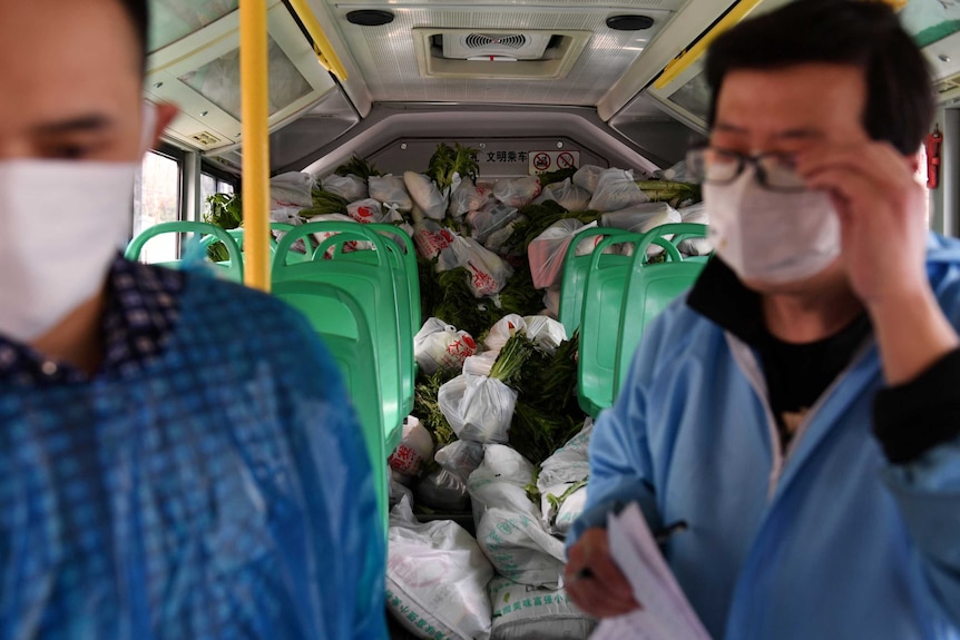 Two people on a bus in face masks with bags of groceries stacked in the back