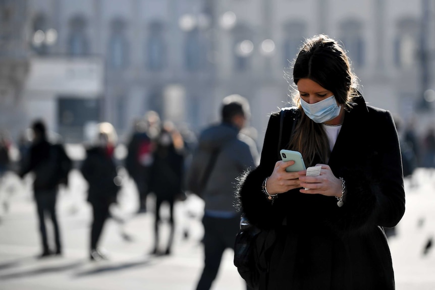 A woman wearing a sanitary mask looks at her phone in Milan.