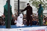 A kneeling woman in all white is hit with a stick by a man dressed completely in black as people watch on and film