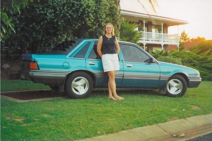 A young woman stands in front of a car in a frontyard