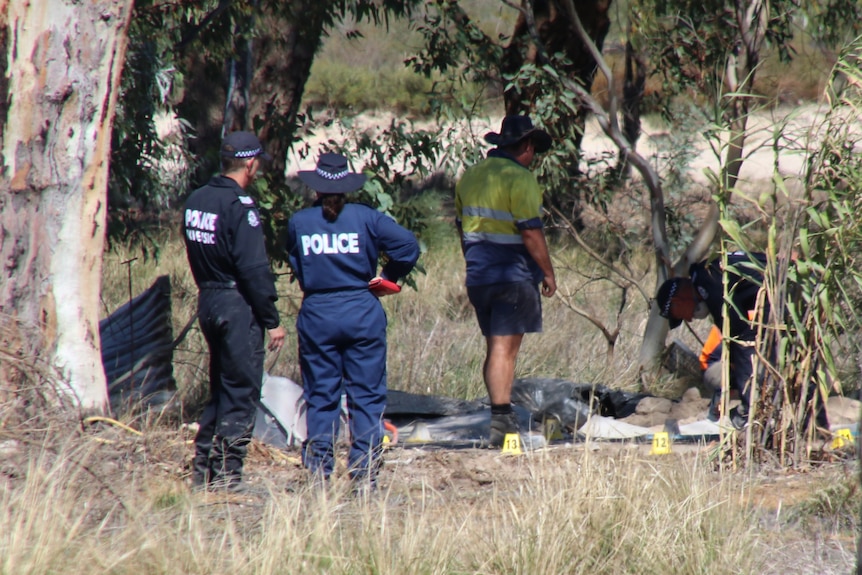 A wide shot showing WA Police forensic officers and a man in a hi-vis vest on a rural property inspecting the ground.