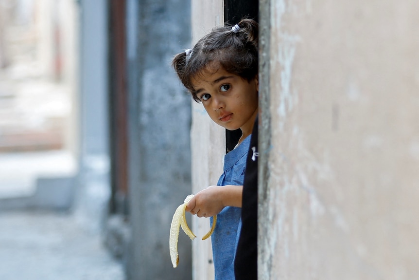 A Palestinian girl holds banana while looking outside the door of her family's house.