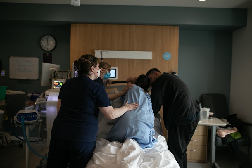 Taryn wearing a blue hospital gown kneeling on a maternity bed, with two midwives and her partner comforting her.