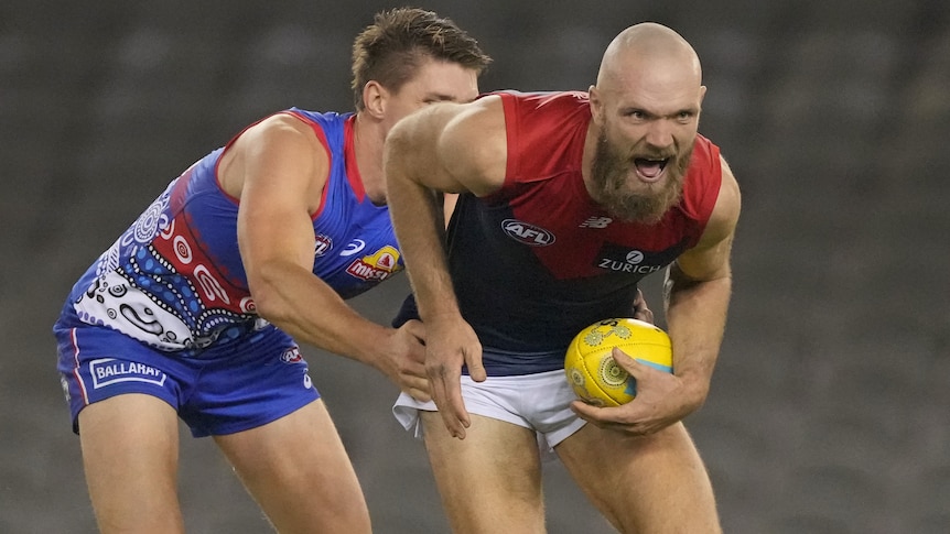 A Melbourne AFL plays holds the ball with his right hand while under defensive pressure from a Bulldogs opponent.