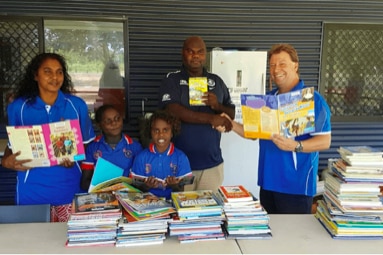 Two Tiwi-Islander kids and three adults stand smiling behind a table loaded with books.