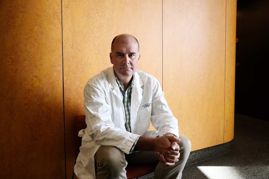 Dr Darren Saunders, in a lab coat, sits in a chair against a pale wooden wall