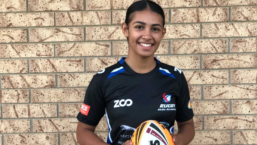 15 Year old girl standing in front of brick wall holding a rugby league ball in a football jersey