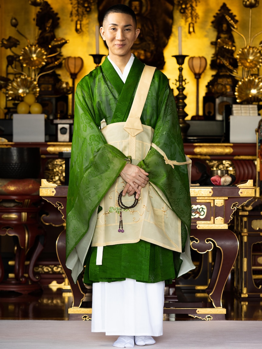 A Buddhist monk in a green gown, surrounded by gold artefacts with a slight smile on his face