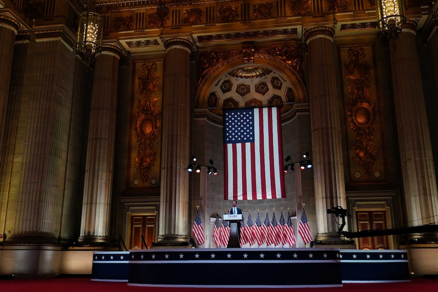 A man stands at a podium on stage with an American flag behind him