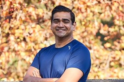 Dulrik Jayasinha in running gear and with wide smile, stands against a brick wall with a leafy tree in the background.