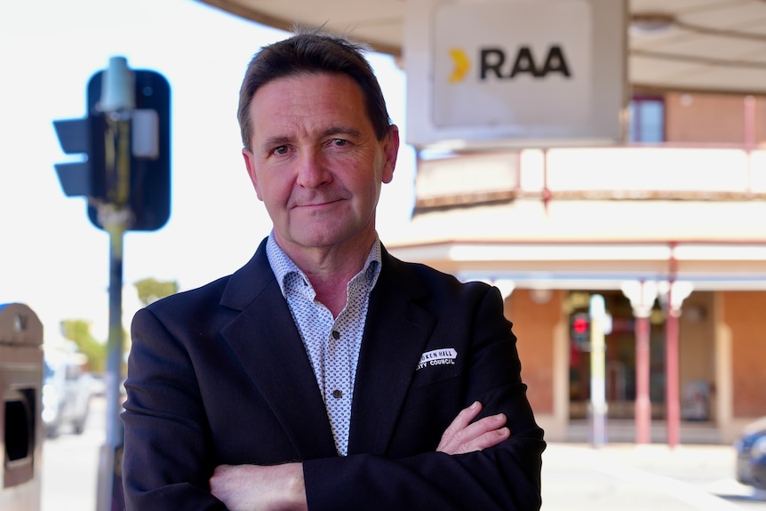A white man with dark hair wearing a black suit and checked shirt crosses his arms in front of the RAA shop in Broken Hill