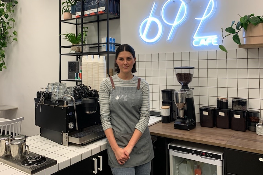 A young woman with short brown hair, wears an apron and stands by a coffee machine in a modern cafe.