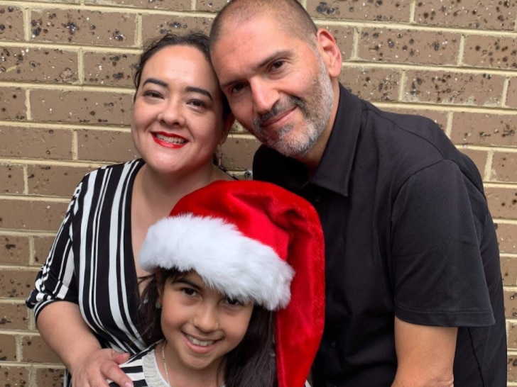 A man, woman and young girl wearing a Santa hat smile as they pose for a family photo in front of a brick wall.