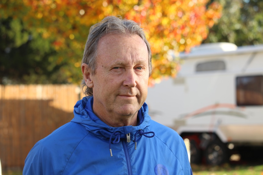 A middle aged man in a blue hoodie stands in front of a caravan