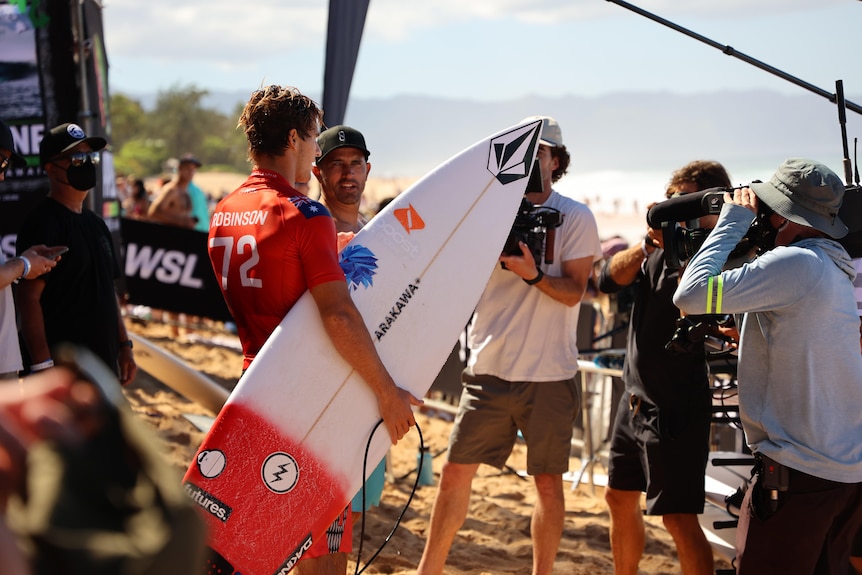 Surfers stand on the beach in front of cameras.