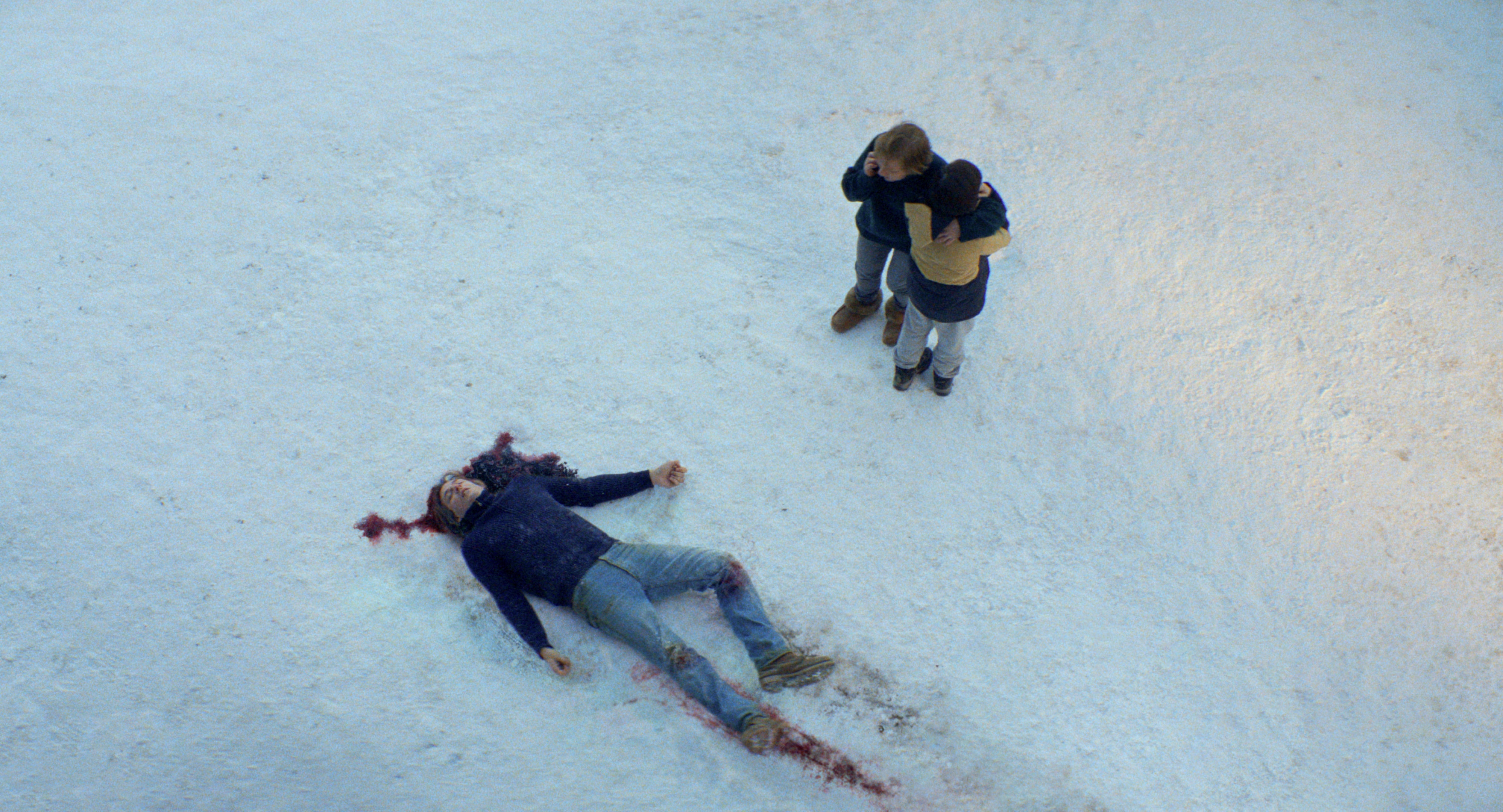 A film still of Samuel Theis as a dead body in blood-stained snow, with Sandra Hüller and Milo Machado Graner hugging nearby.