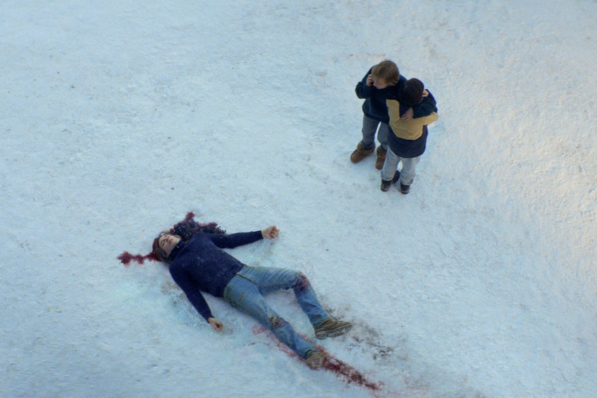 A film still of Samuel Theis as a dead body in blood-stained snow, with Sandra Hüller and Milo Machado Graner hugging nearby.