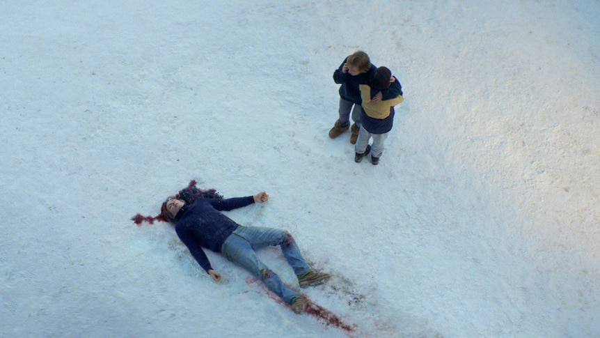 A film still of Samuel Theis as a dead body in blood-stained snow, with Sandra Hüller and Milo Machado Graner hugging nearby.