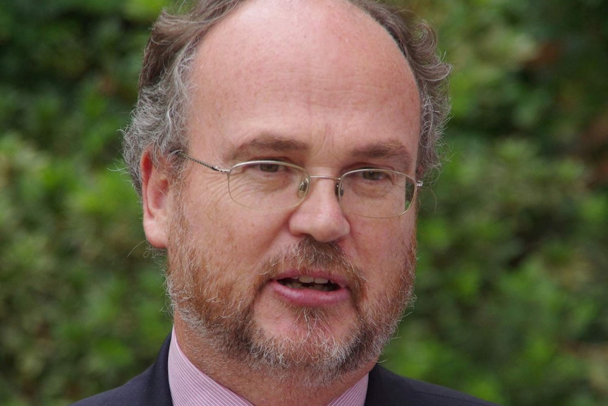 Bespectacled man wearing suit and tie, with greenery in the background