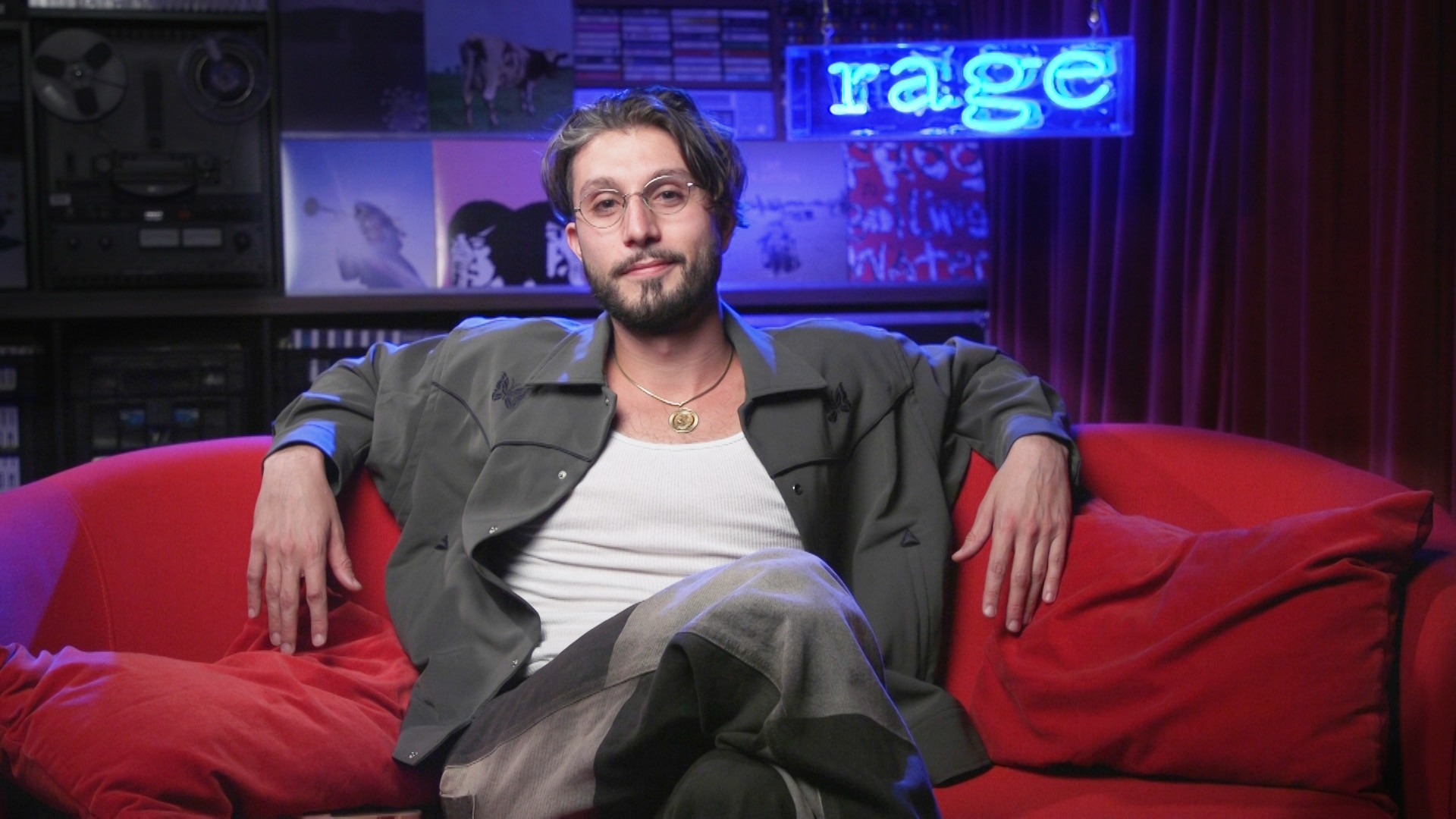 baby no money sits on a red couch in front of a blue neon sign that reads rage