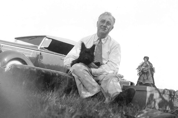 Black and white photo of FDR sitting outside next to a car with a small black Scottish terrier puppy on his lap.