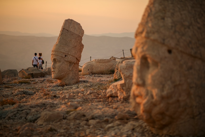Üç kişilik bir aile, dev kafaların taş heykelleriyle çevrili kayalık bir manzaraya bakmaktadır.