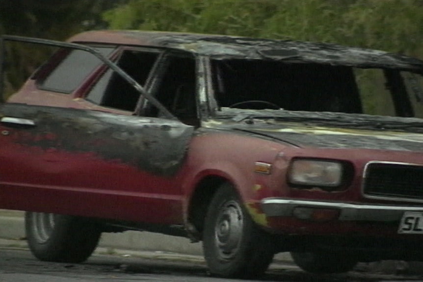 A red burnt-out car. One if its doors is open and the windscreen is gone.