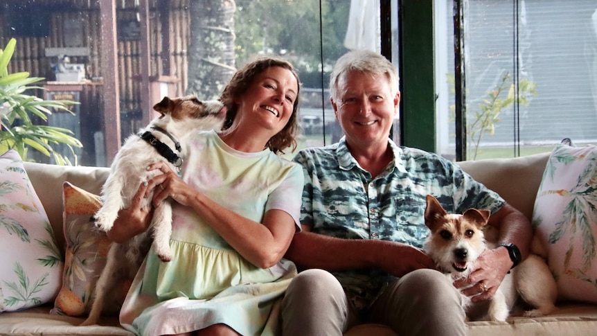 Middle-aged woman and man sitting on a couch smiling at the camera, petting two dogs.