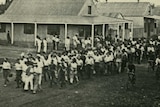 Black and white photo of an old car followed by a group of people marching down a street in 1918.