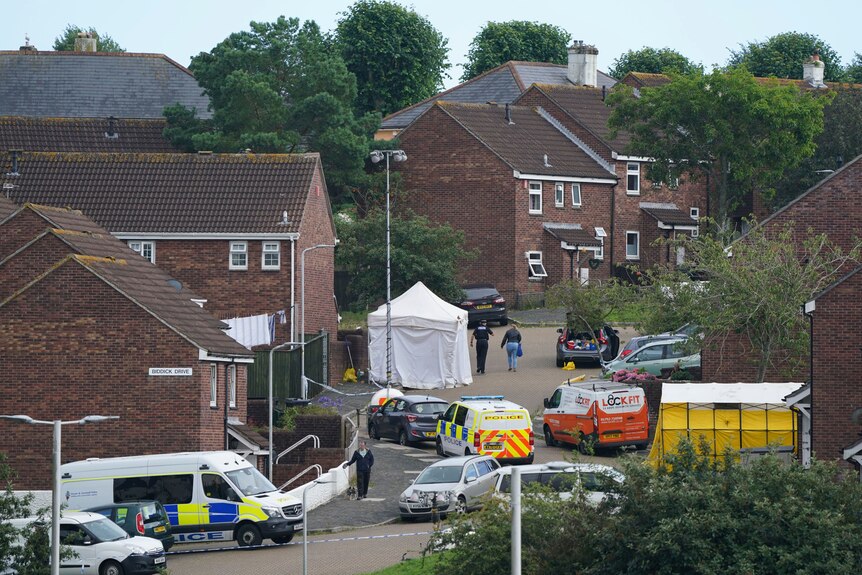 Forensics teams and ambulances in a small street with dark-brown brick houses.