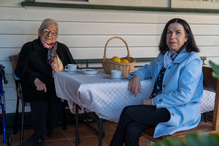 an elderly woman and her daughter in her 60s sit at an outdoor table having cups of tea. 