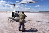 A helicopter sits on a salt lake while a man makes a sample hole with a hand auger.