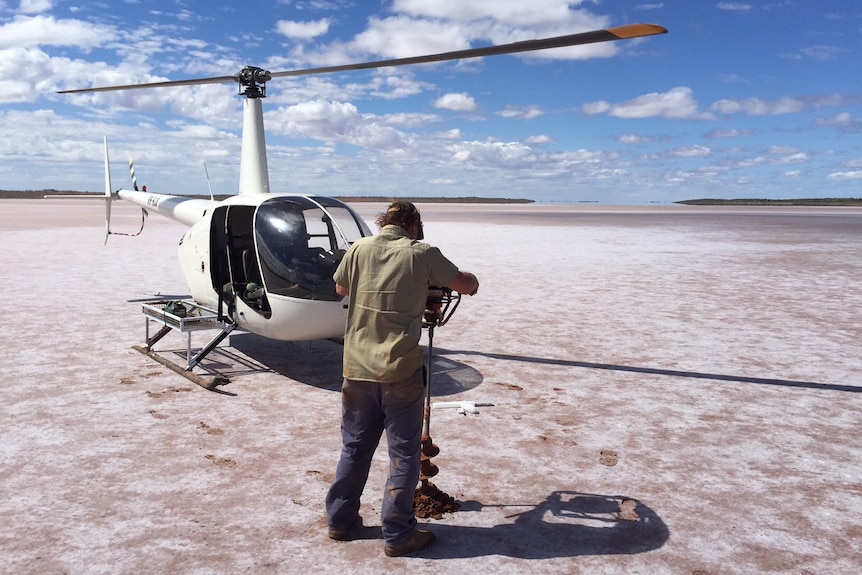A helicopter sits on a salt lake while a man makes a sample hole with a hand auger.
