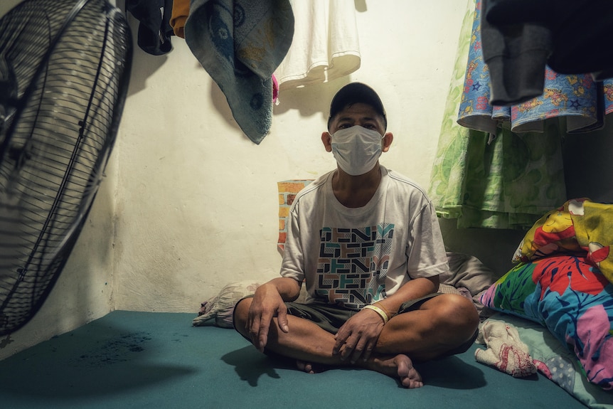 A Filipino man in a white face mask and cap sits on the floor of a house 