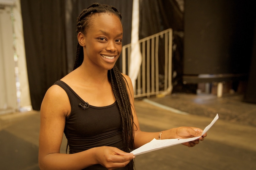 A younng woman holding a piece of paper, smiles at the camera. There is a small microphone pinned to her singlet.