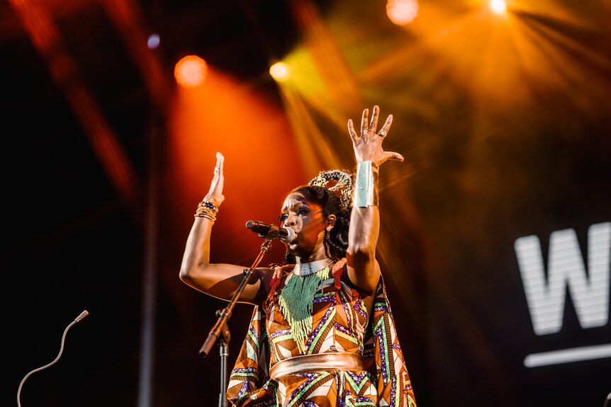 A woman in a dress with African print and a headress holds her arms aloft