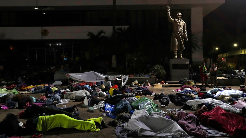 Dozens of people sleep on the ground in a public square at the base of a statue of a man with his arm raised in the air