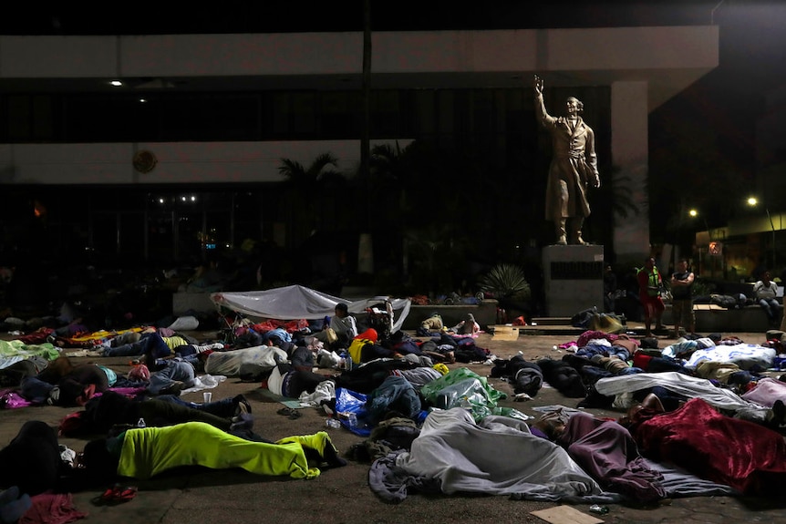 Dozens of people sleep on the ground in a public square at the base of a statue of a man with his arm raised in the air