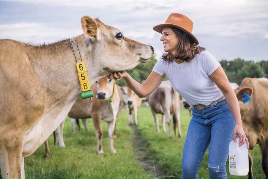 Woman with short brown hair wearing a brown hat, white shirt and jeans lovingly scratching under a cow's chin in cow paddock.