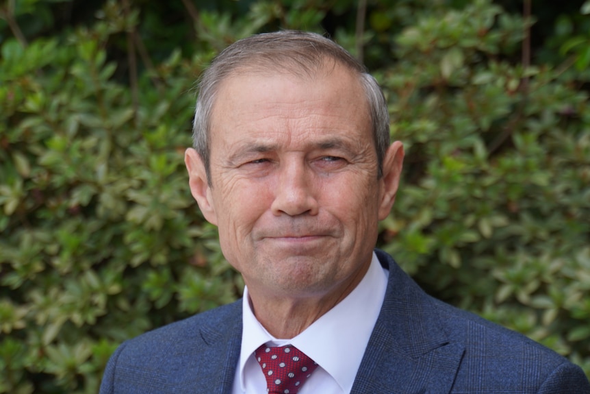 A  head and shoulders shot of Roger Cook outdoors at a media conference, wearing a suit and tie.