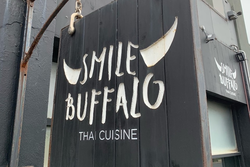 A black sign with 'Smile Buffalo Thai Cuisine' written on it hangs outside a restaurant.