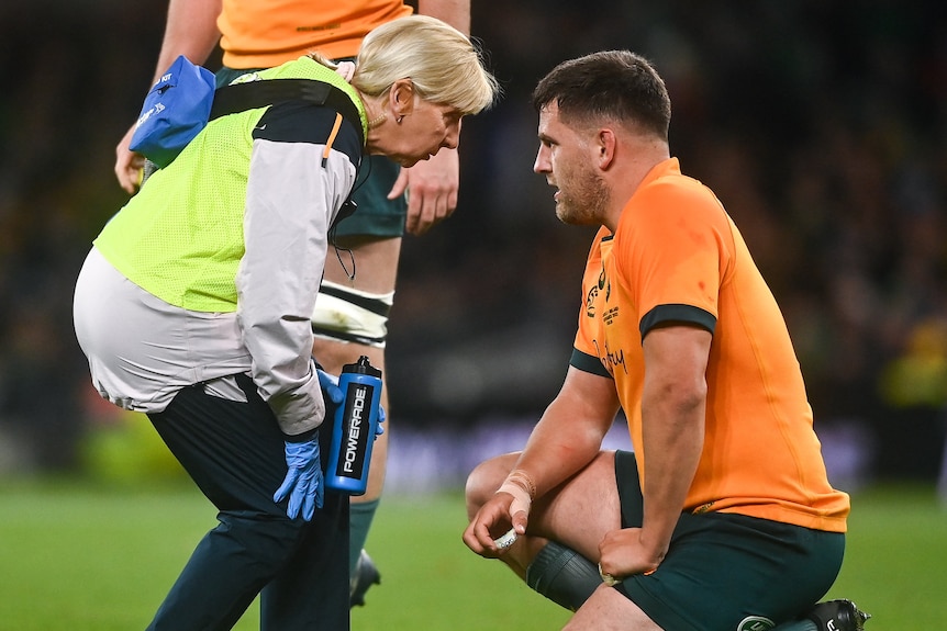An Australian rugby player kneels with his mouthguard in one hand as a doctor assesses him on the pitch. 