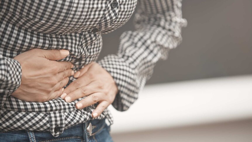A close up of a woman wearing a gingham shirt and jeans clutching stomach as if in pain.