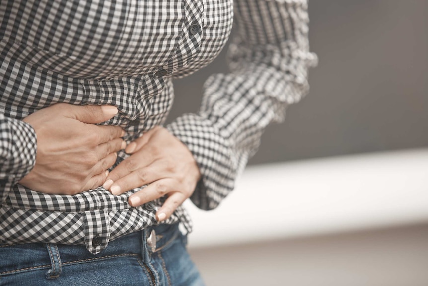 A close up of a woman wearing a gingham shirt and jeans clutching stomach as if in pain.