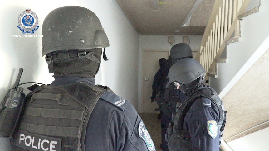 four men wearing dark helmets with coverings over their mouths stand outside a door