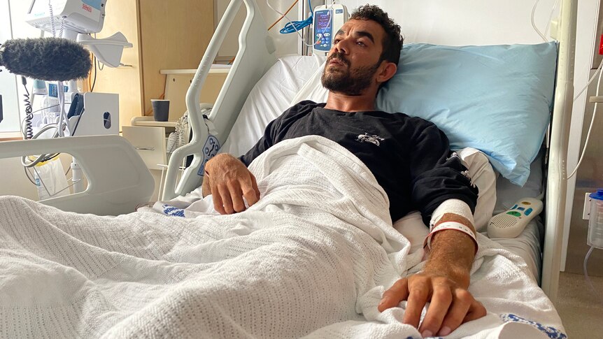 A young Indigenous man lies in a hospital bed recovering from a shark attack.
